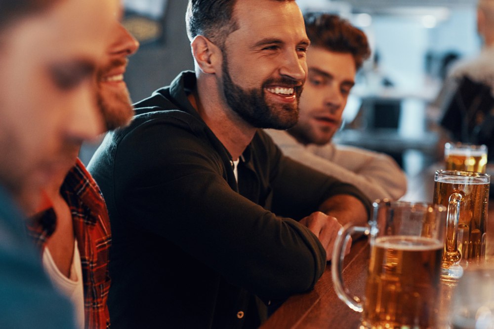 Jeune homme heureux en tenue décontractée dégustant une bière et souriant alors qu'il est assis au pub.